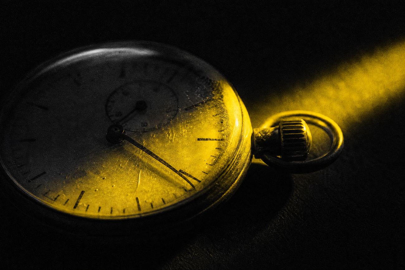 Close-up of an old analogue stopwatch on a dark surface, one narrow yellow beam of light across the dial